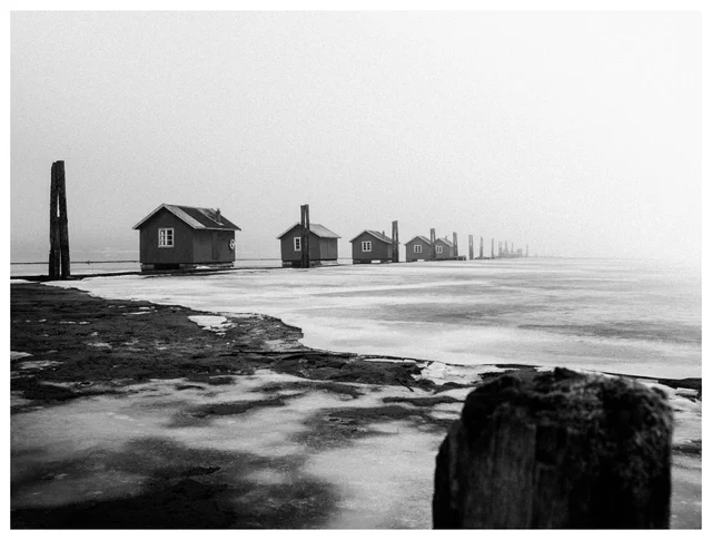 Row of small cabins and wooden poles fading into fog along a frozen shoreline in winter, minimalist black and white landscape.
