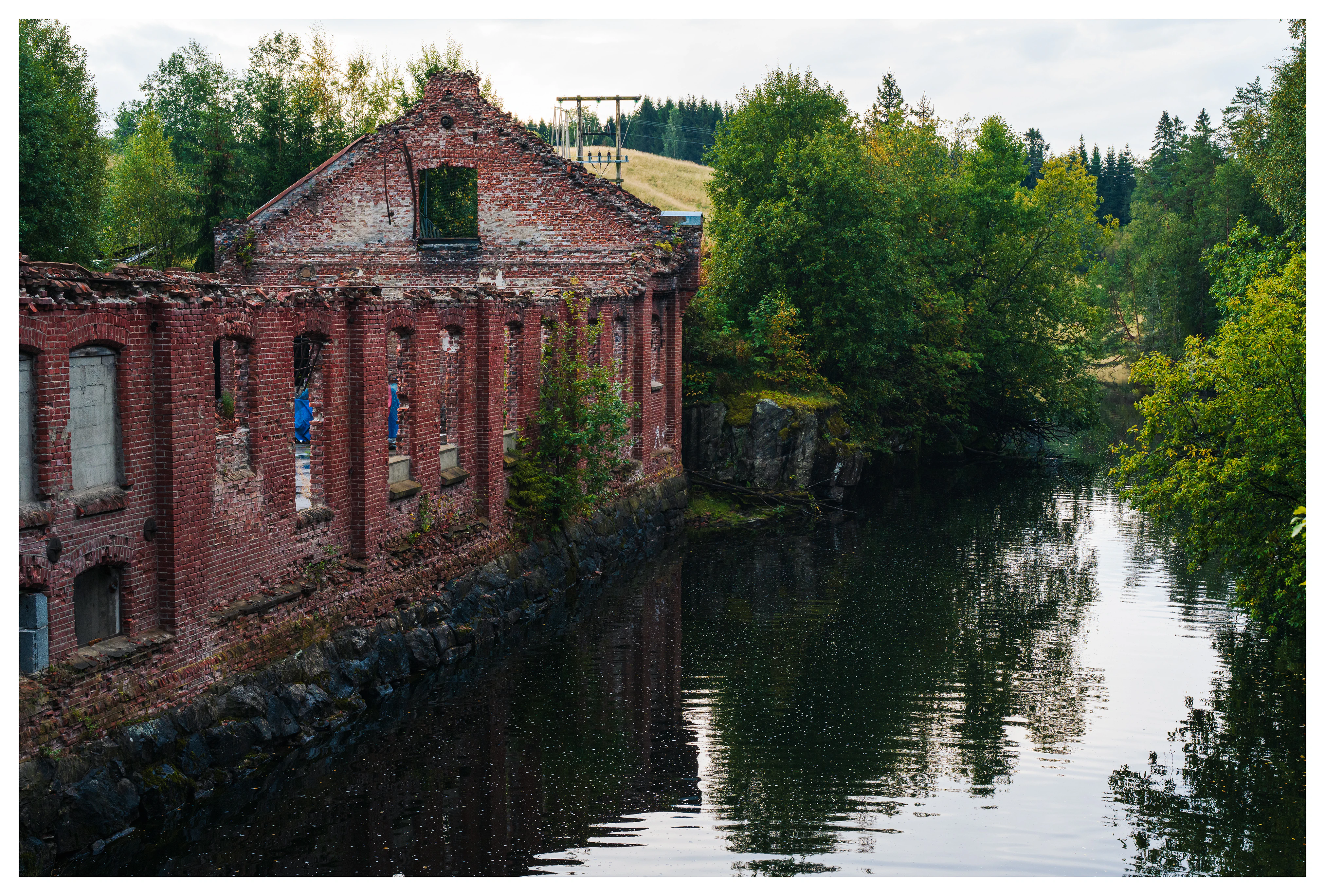 Abandoned brick walls of the old Mago C power plant standing beside the calm Andelva river in Eidsvoll, surrounded by dense green forest and reflected water.