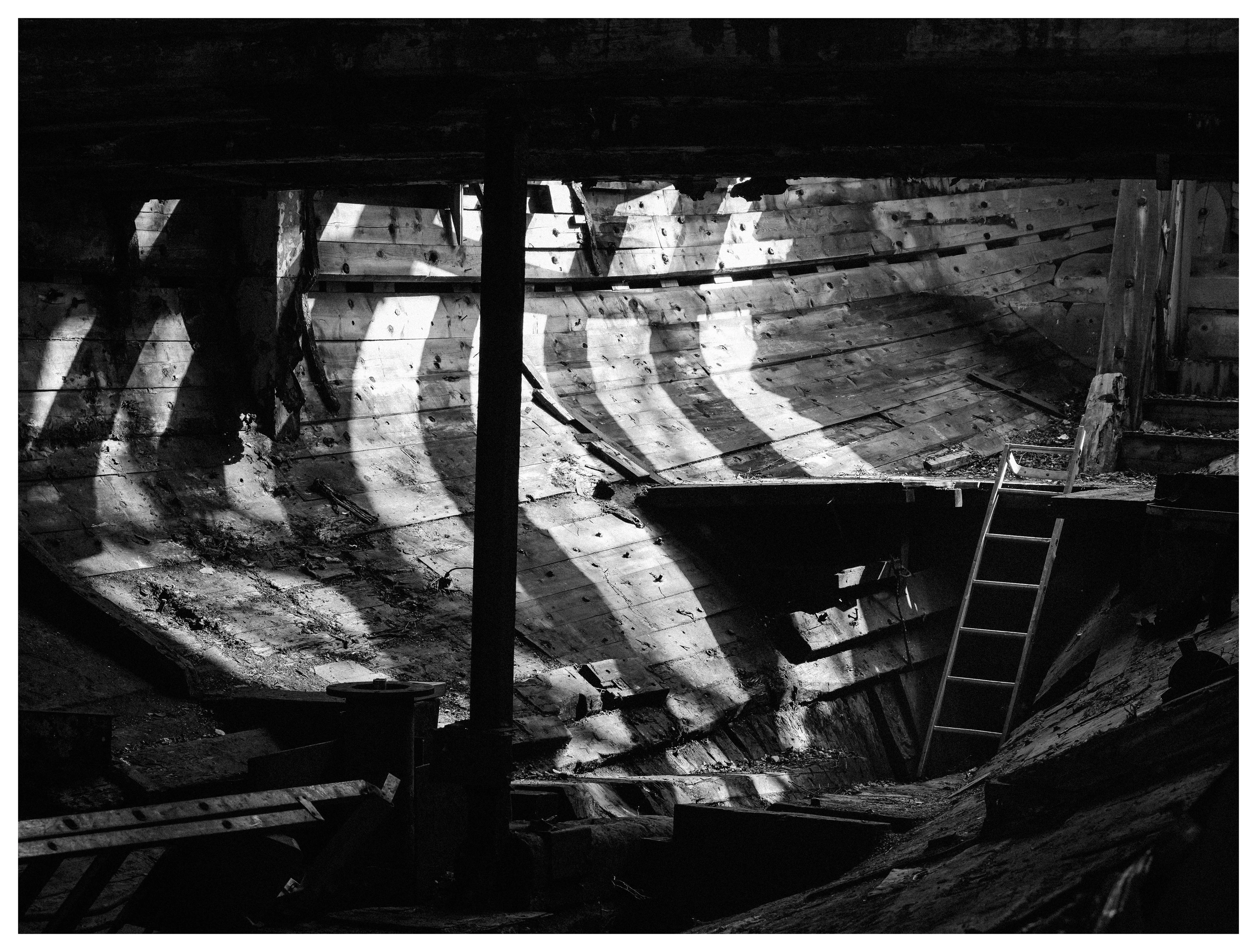Black and white photo of an old wooden boat interior with curved hull, ladder, and light streaming through beams.