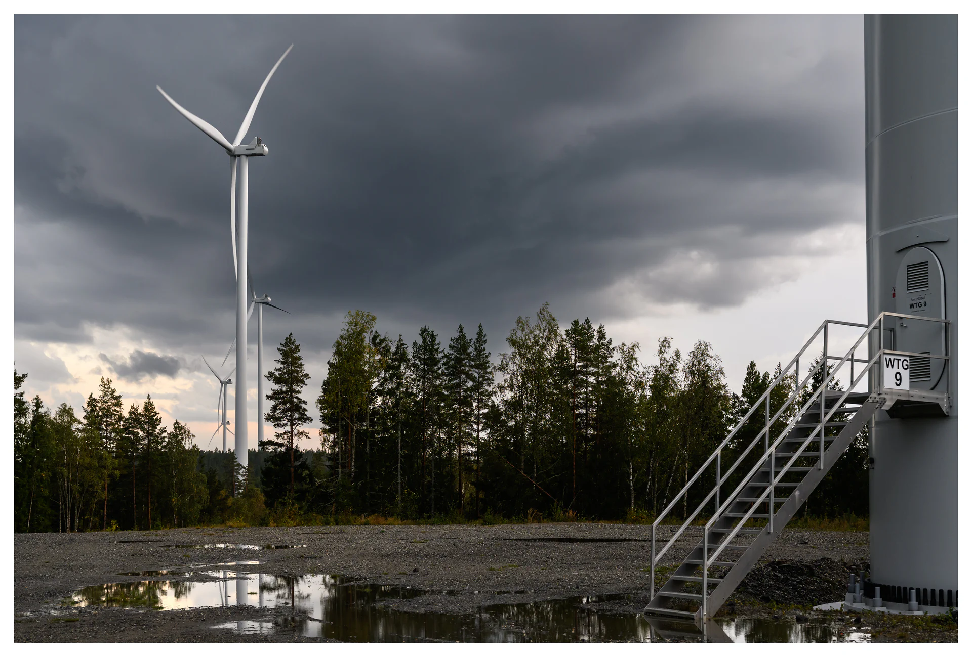 Color photo of wind turbines and a turbine access staircase at the Marker wind farm in Norway, with puddles, forest trees and dramatic storm clouds.