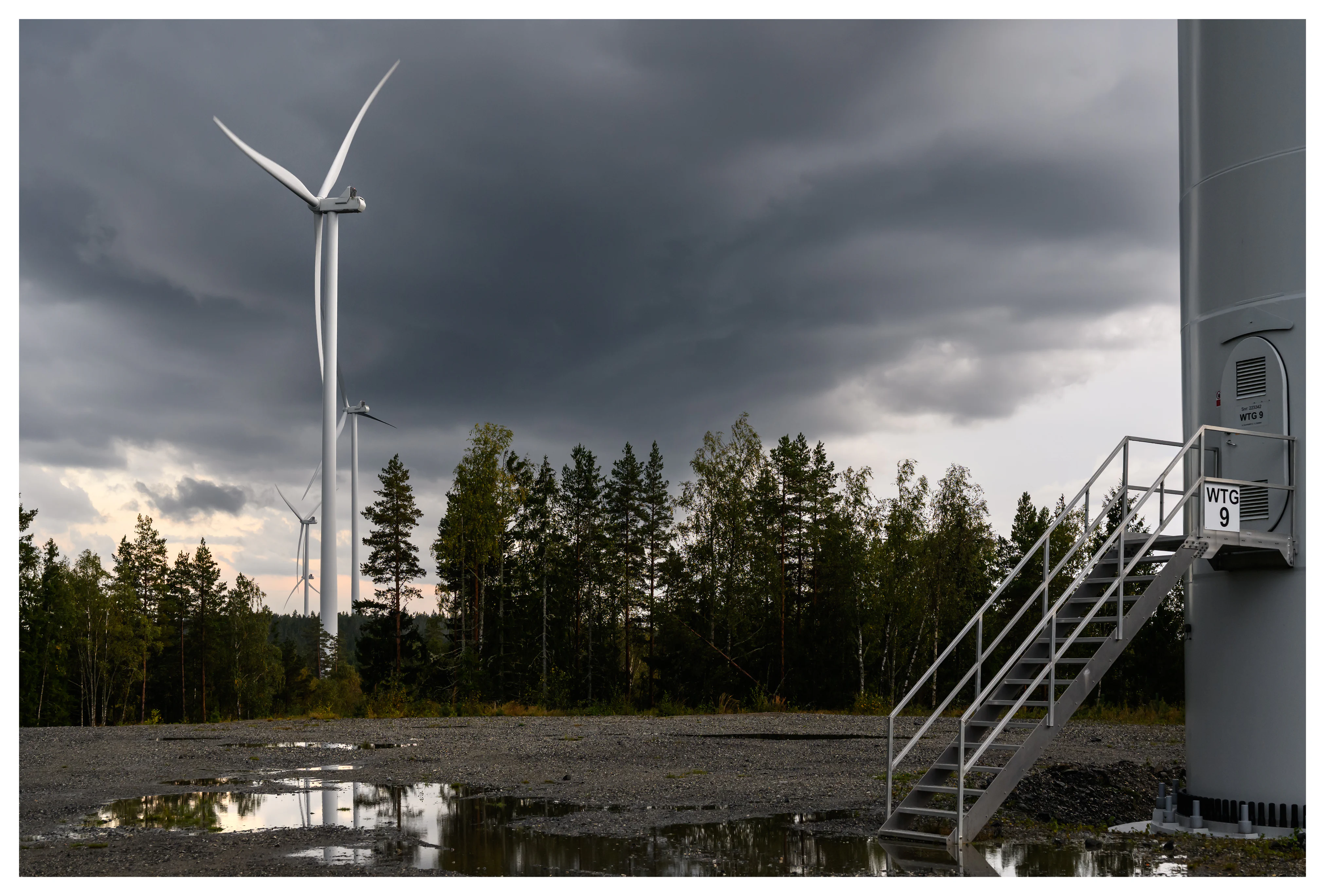Color photo of wind turbines and a turbine access staircase at the Marker wind farm in Norway, with puddles, forest trees and dramatic storm clouds.