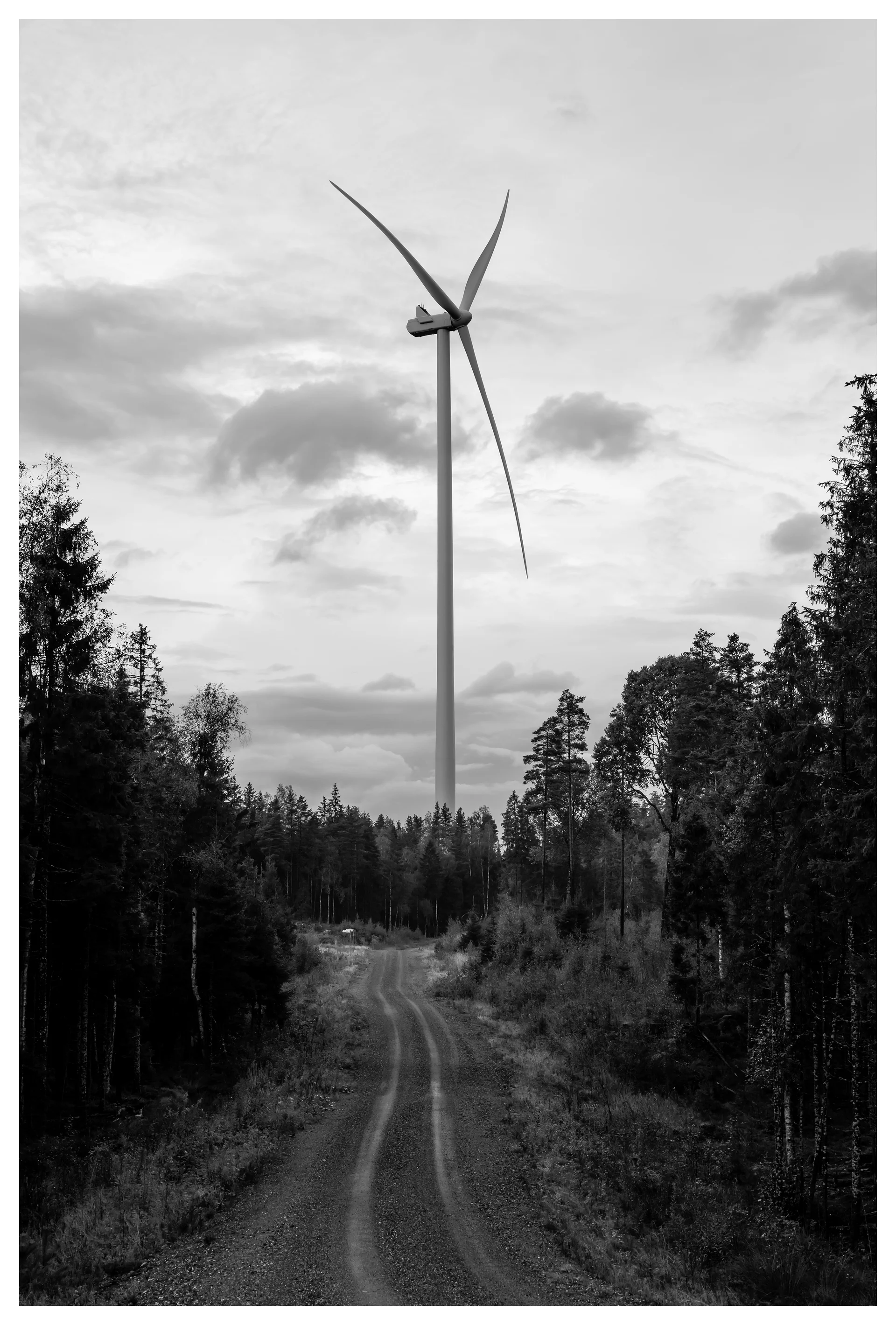 Black and white photo of a tall wind turbine at the end of a gravel road in the Marker wind farm, Norway, surrounded by dense forest and cloudy sky.