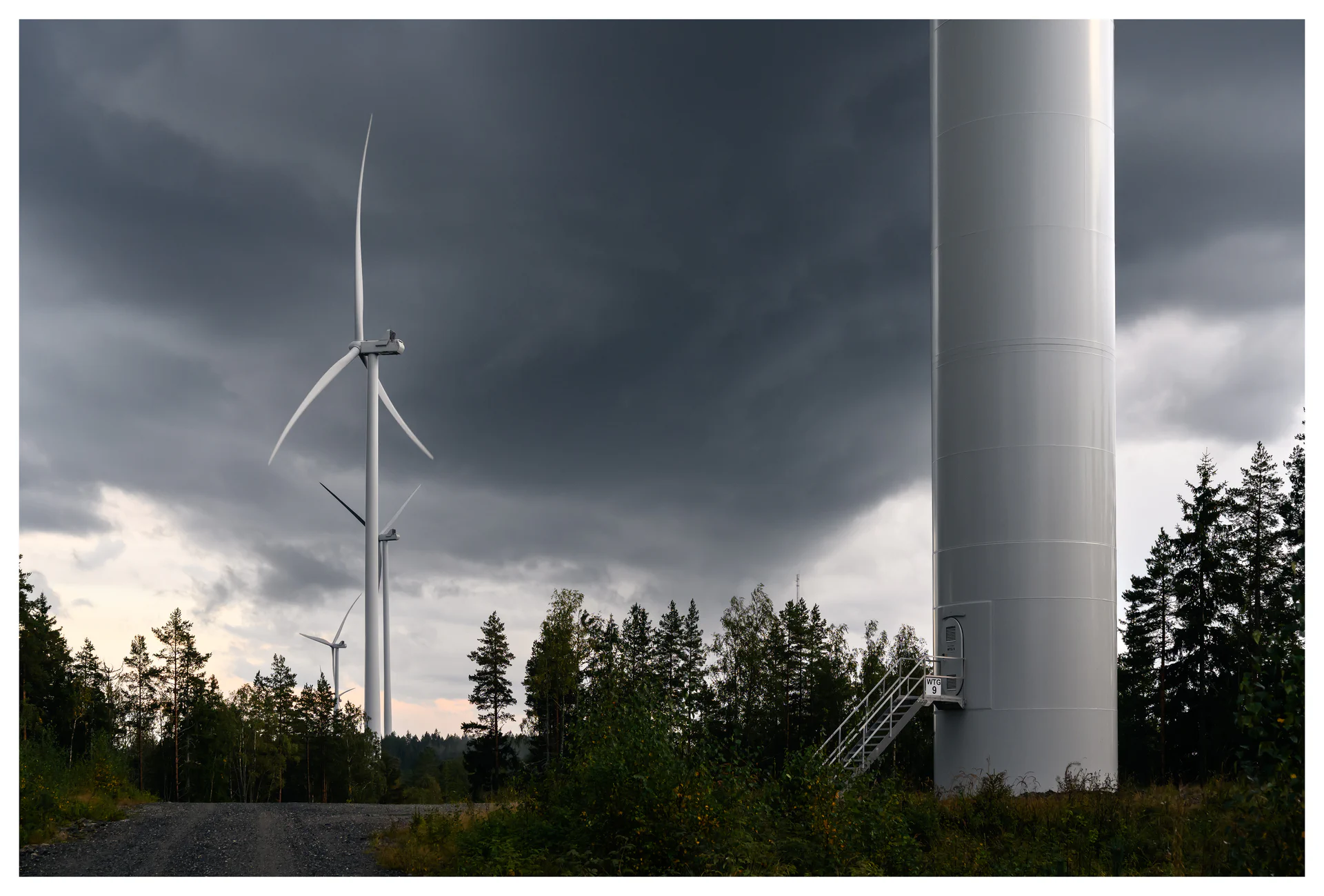 Color photo of wind turbines at the Marker wind farm in Norway, with a turbine tower and forest vegetation in the foreground under dark storm clouds.