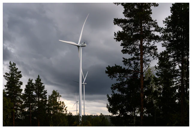 Color photo of wind turbines at the Marker wind farm in Norway, surrounded by tall forest trees under dark storm clouds and dramatic light.