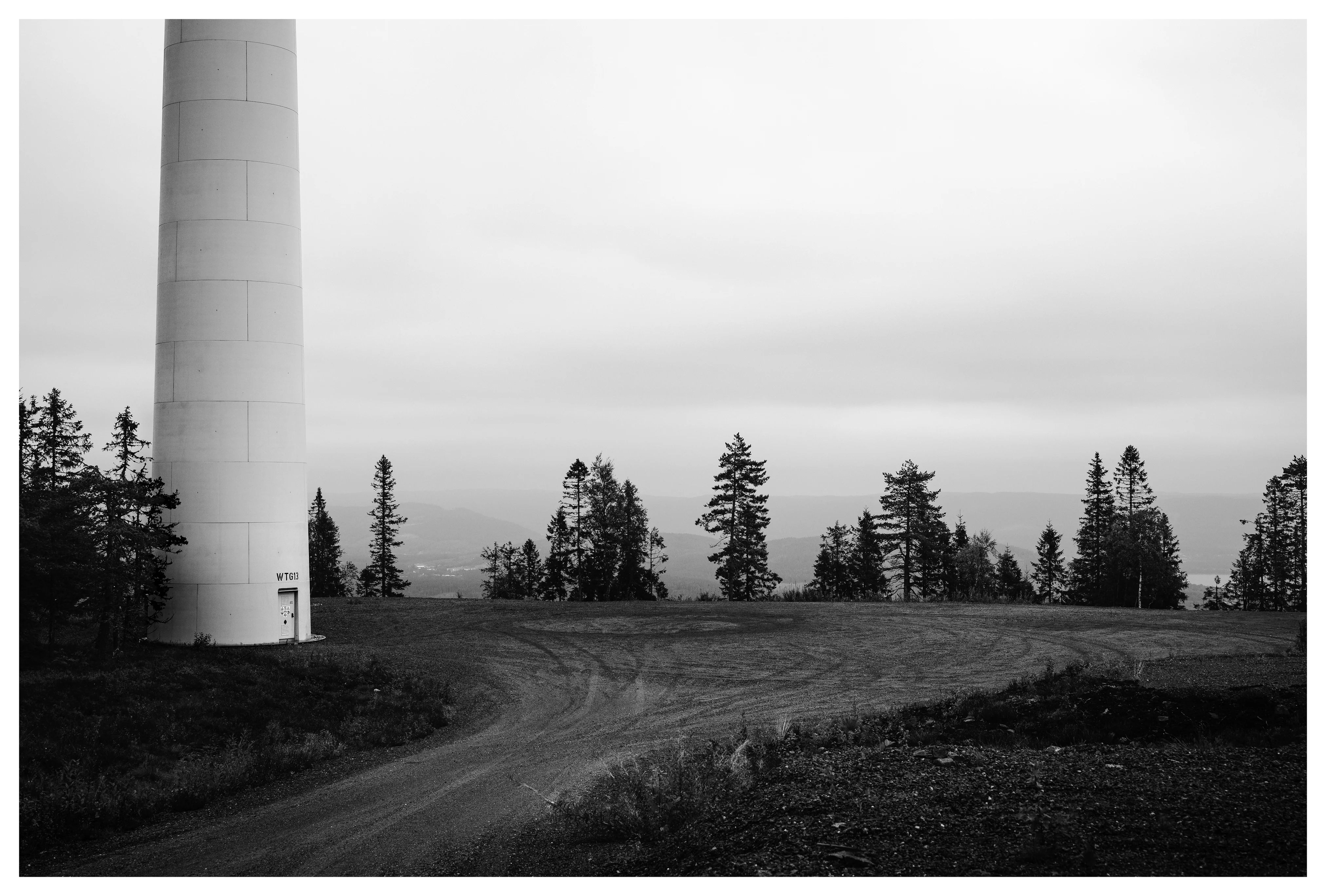 Black and white photo of a wind turbine base at Odal wind farm in Norway, with forest trees, gravel road and distant hills under cloudy sky.