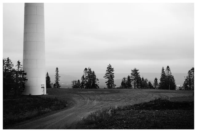 Black and white photo of a wind turbine base at Odal wind farm in Norway, with forest trees, gravel road and distant hills under cloudy sky.