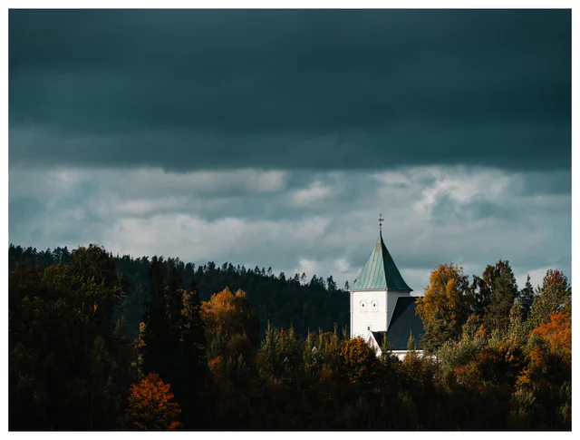 White church surrounded by autumn trees beneath dark gathering storm clouds in the Norwegian countryside.